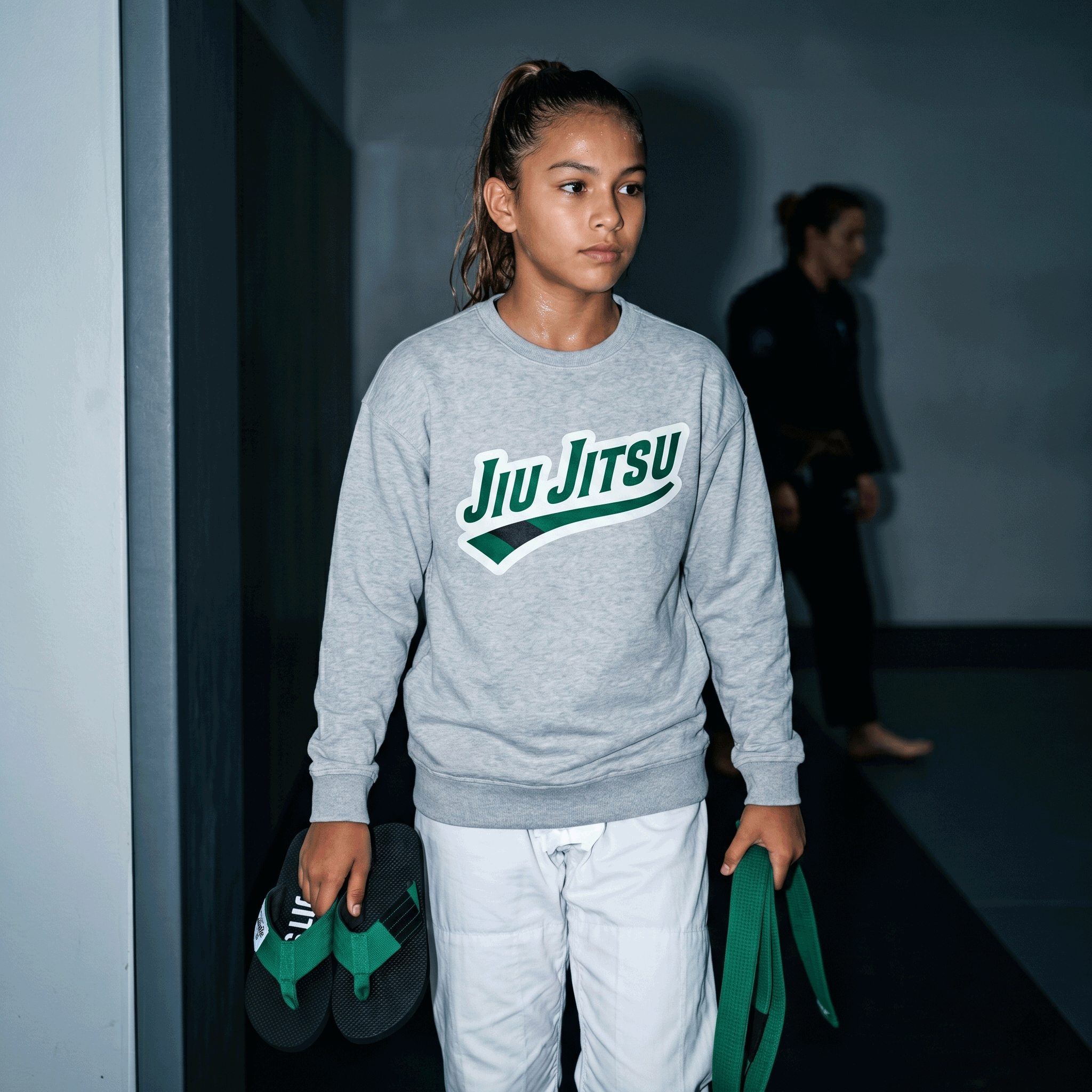 Young BJJ practitioner wearing FlowFlip Co. sweatshirt and carrying green belt flip flops and a green belt in the academy hallway